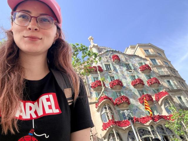 AmbeerWood with glasses, red cap, and black Marvel shirt, standing in front of a colorful building with red flower boxes. Bright blue sky, sunny day.