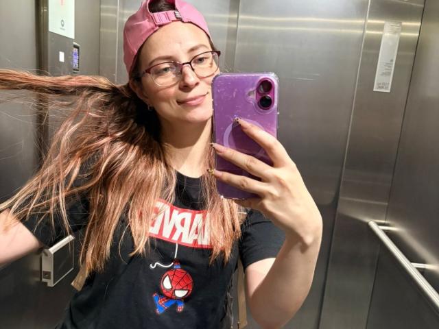AmbeerWood, wearing glasses, pink cap, and Spider-Man shirt, holds phone with purple case, smiling, in elevator, with long brown hair flying. AmbeerWood has medium tits.