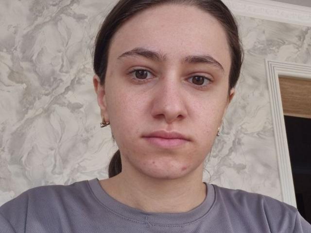SherilDerico hat ein ernstes Gesicht, braune Augen und helle Haut mit einigen Pickeln am Kinn. Sie trägt ein graues Hemd und kleine Ohrringe, und ihr braunes Haar ist zurückgebunden..
