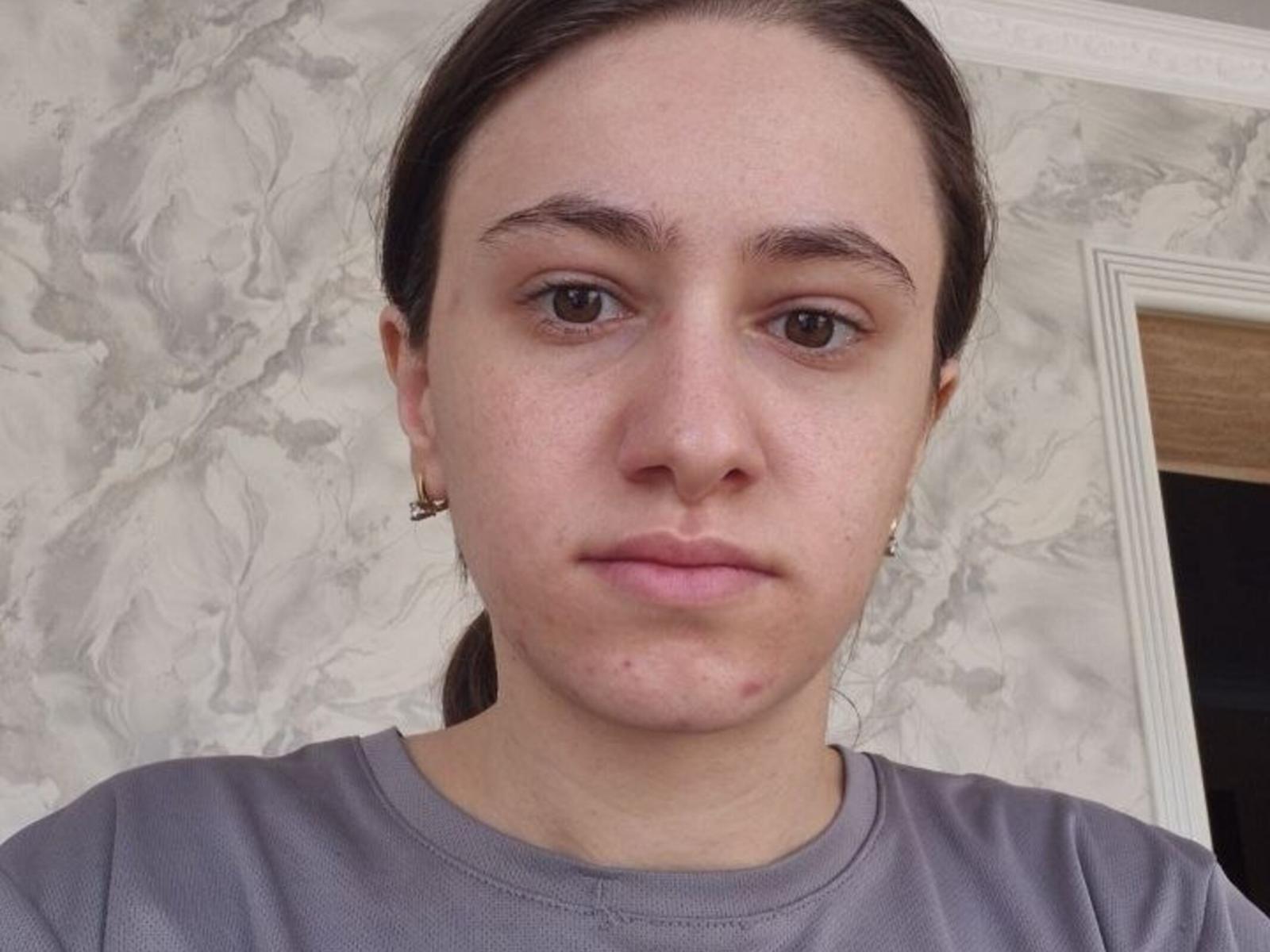 SherilDerico tiene una expresión seria, ojos marrones y piel clara con algunos granos en la barbilla. Lleva una camisa gris y pequeños aretes, con el cabello castaño recogido..
