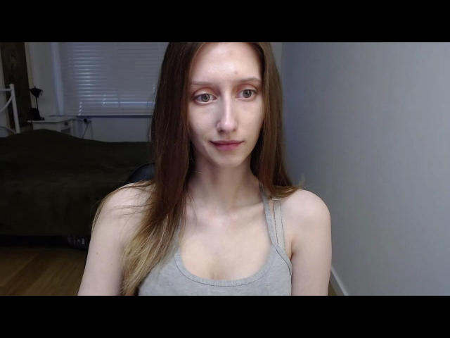 LunaXKiss hat langes, braunes Haar, blasse Haut, kleine Brüste und ein leichtes Lächeln. Sie trägt ein graues Tanktop und steht in einem Raum mit einem grünen Bett..