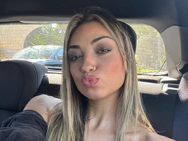 CameliaDeihl with pouty pink lips, light brown eyes, and blonde hair, sitting in a car, wearing a black shirt, showing her nose ring, and looking to the side.