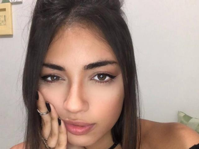 CameliaDeihl has tan skin, brown eyes, dark eyebrows, long black hair, and is touching her cheek with a hand showing a ring, looking slightly pouty with pink lips.