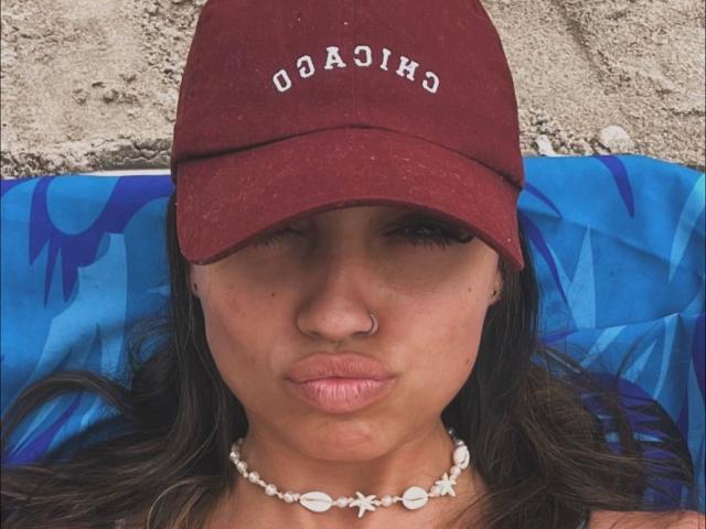 CameliaDeihl has brown hair, a nose ring, and a red cap with "COAST" in white. She's pouting with pink lips, wearing a white shell necklace, lying on a blue towel.