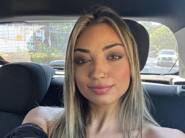 CameliaDeihl has light brown skin, straight blonde hair, dark eyebrows, green eyeshadow, pink lips, and a nose ring, sitting in a car with black headrests.