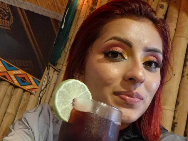 LizBonnet, con cabello rojo, sombra de ojos amarilla y labial, sosteniendo un vaso con un líquido oscuro y lima, fondo de pared con paneles de madera, piel ligeramente brillante, con una sonrisa burlona..