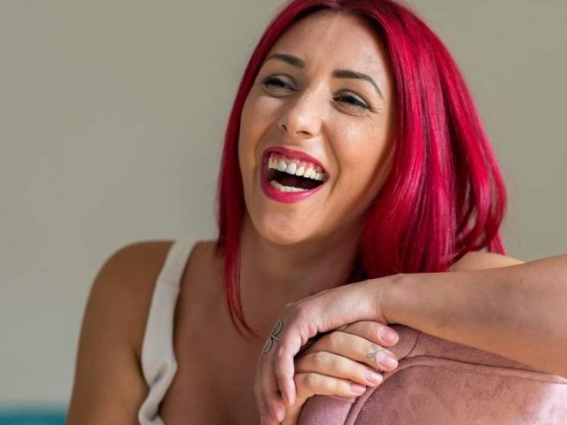 BubbleButtAlice, con i capelli rosso acceso, sorride ampiamente con la bocca aperta, mostrando i denti. Ha una mano appoggiata su un cuscino rosa e indossa una maglietta bianca..