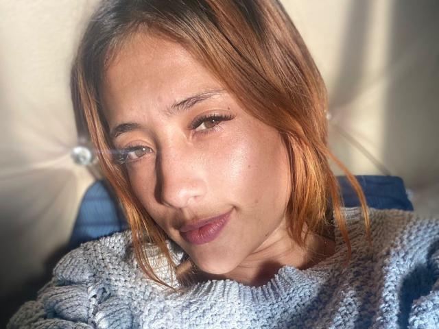 ZoeTaylorl with light brown, slightly wavy hair, wearing a knitted gray sweater, has a subtle smile and sunlit, slightly shiny face, showing small nose and brown eyes.