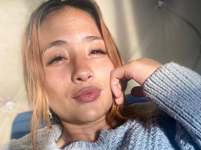 ZoeTaylorl with pouty lips, light brown hair, and a gray knitted sweater, holding her cheek, sunlight on her face, slightly squinting eyes.