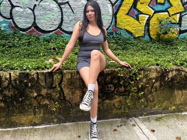 MaraCortes, with dark hair, gray tank top, and shorts, sits on a concrete wall, one leg raised, wearing white socks and black sneakers, showing her ass.