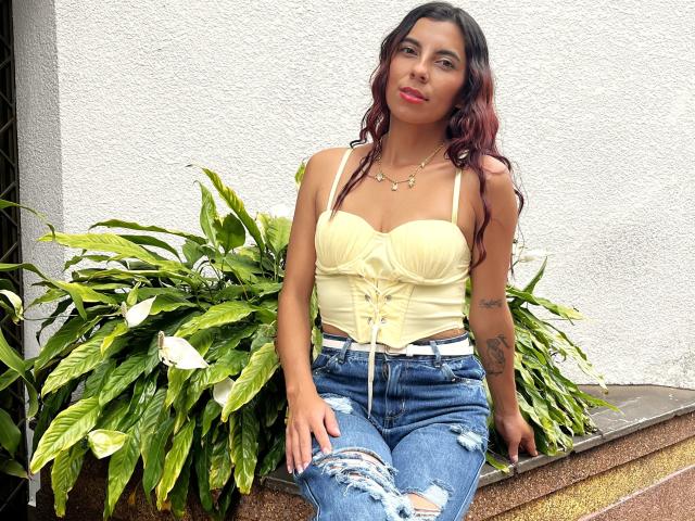 Carla Torres, met een medium bruine huid, golvend donkerrood haar, een gele top zonder bandjes, waarbij haar kleine borsten zichtbaar zijn, versleten blauwe jeans, tatoeages op haar rechterarm, zittend naast een groene plant, met een glimlach..