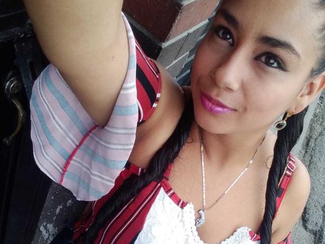 AlisonHank with pink lipstick, black braids, red striped dress, white lace, silver necklace, and earrings, holding up a selfie. Boobs visible in dress. Smiling, pink lips, dark eyes.