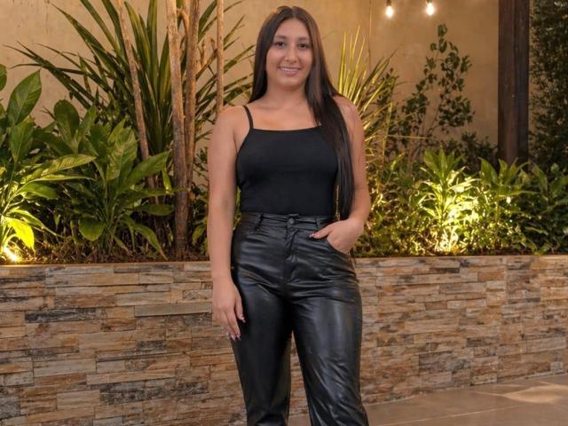 Catalina Rivero se tient debout, avec de longs cheveux bruns, vêtue d'un débardeur noir et d'un pantalon en cuir noir brillant, souriante, une main dans la poche, avec des plantes vertes luxuriantes et un mur de pierres empilées en arrière-plan..