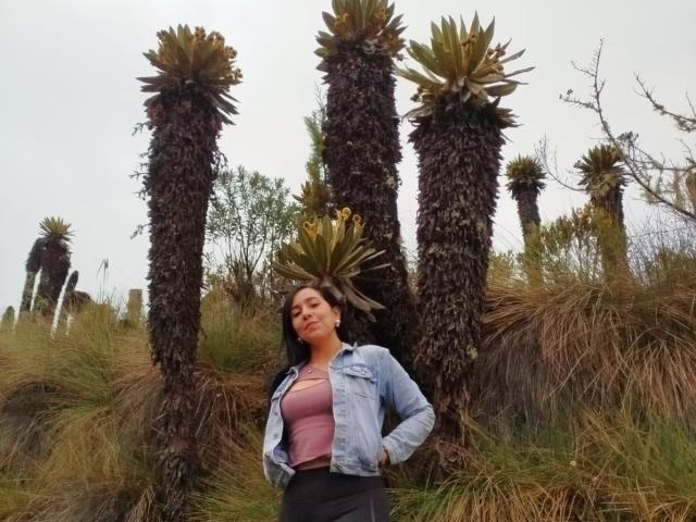 KattyRivera stands in front of tall palm trees with spiky tops, wearing a denim jacket and pink top, hands in pockets, looking slightly to the side with a relaxed expression.