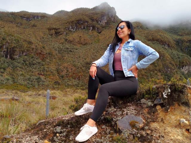 KattyRivera with medium tits, wearing a denim jacket, black leggings, and white sneakers, sitting on a rocky outcrop, smiling, with fog-covered mountains in the background.