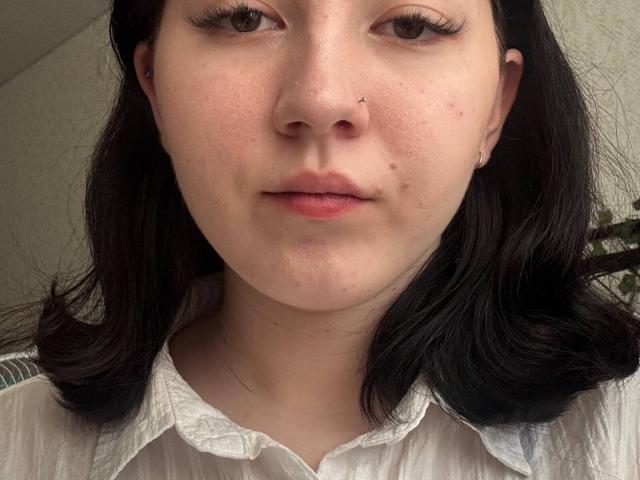 CorneliaCarrethers has a pale skin tone with slight acne, black shoulder-length hair, a small nose piercing, and wears a white button-up shirt. Her eyes are brown and she's looking forward.
