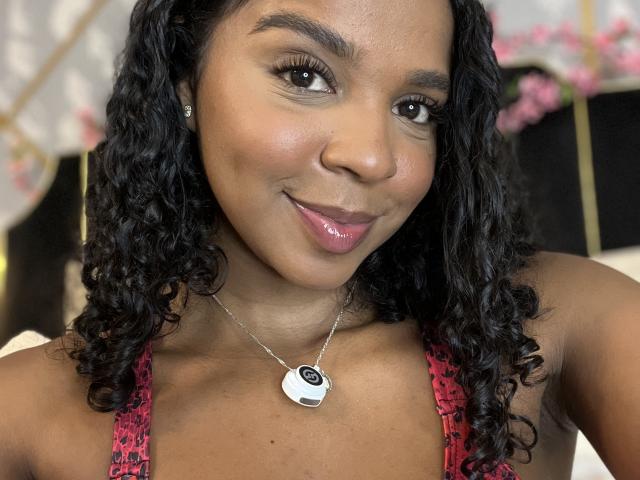 LisaMendez has dark curly hair, brown skin, and a shiny pink lip. She's wearing a red lace top and a silver necklace with a black and white pendant.