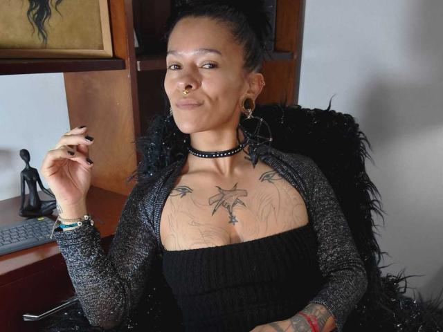 AmelieBlake with tattoos on her chest and arms, wearing a black choker and sheer gray cardigan, sitting in a chair, smiling with a nose ring, and black nail polish.