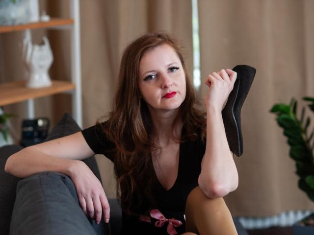 MeganPirs with red lipstick, holding black high heel in right hand, leg raised, wearing black top, pink ribbon, brown hair, sitting on gray couch, looking to side.