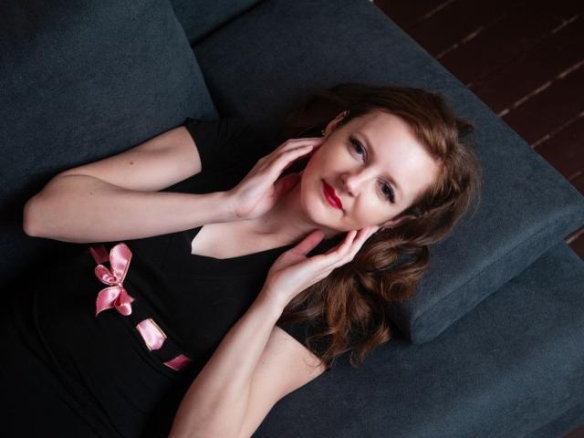 MeganPirs with red lipstick, brown wavy hair, black dress with pink ribbon, hands near face, lying on dark gray couch, smiling subtly, pale skin, feminine, relaxed.