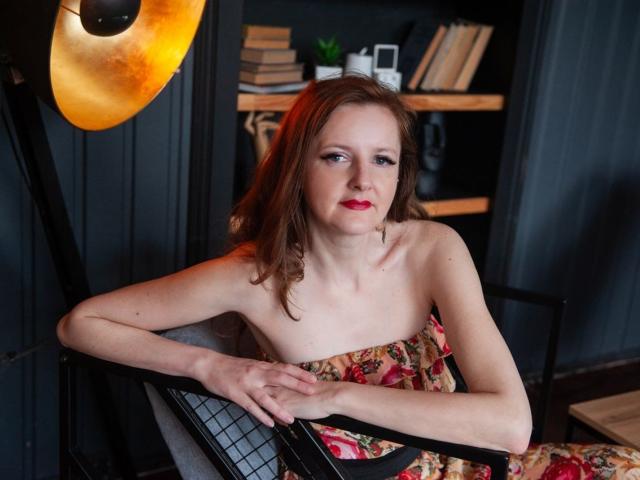 MeganPirs with red hair, sitting, arms crossed, wearing a floral dress, showing her small tits, under a warm light, looking to the side with red lipstick.