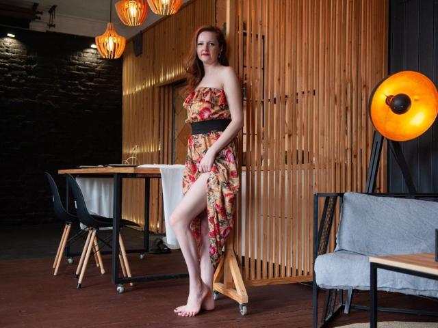 MeganPirs with red hair, wearing a floral strapless dress, standing barefoot, showing her ass, in a room with wooden panels, black chair, and orange hanging lights.