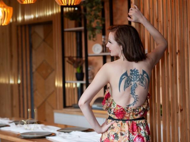 MeganPirs with a dragon tattoo on her back, wearing a floral dress, standing with one arm raised, showing her bare back and tits, in a wooden room.