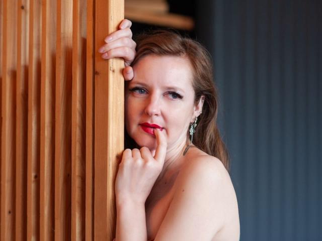 MeganPirs with red lipstick, brown hair, and earrings, peeks from wooden slats, finger to mouth, showing bare shoulders, soft smile, sexy, looking at camera.
