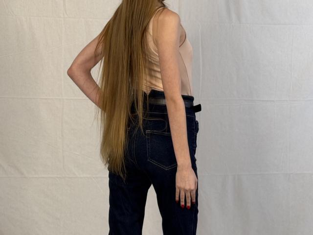 YangDeschenes, con lunghi capelli biondi, indossa una canotta beige e jeans blu scuro, è in piedi con la schiena rivolta verso la fotocamera, una mano appoggiata al fianco, mostrando i glutei..
