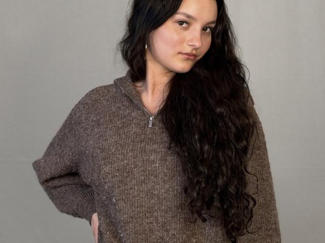 MegganCarlson with long wavy black hair, wearing a brown knit sweater, hands in pockets, zipper partially down, slight smirk, gray background. Boobs not visible.