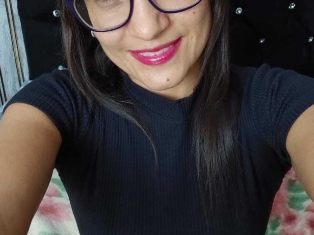 SolaraDubois with dark hair, glasses, and pink lipstick, smiling at the camera, wearing a black ribbed shirt, showing her chin and upper chest. Boobs not visible.