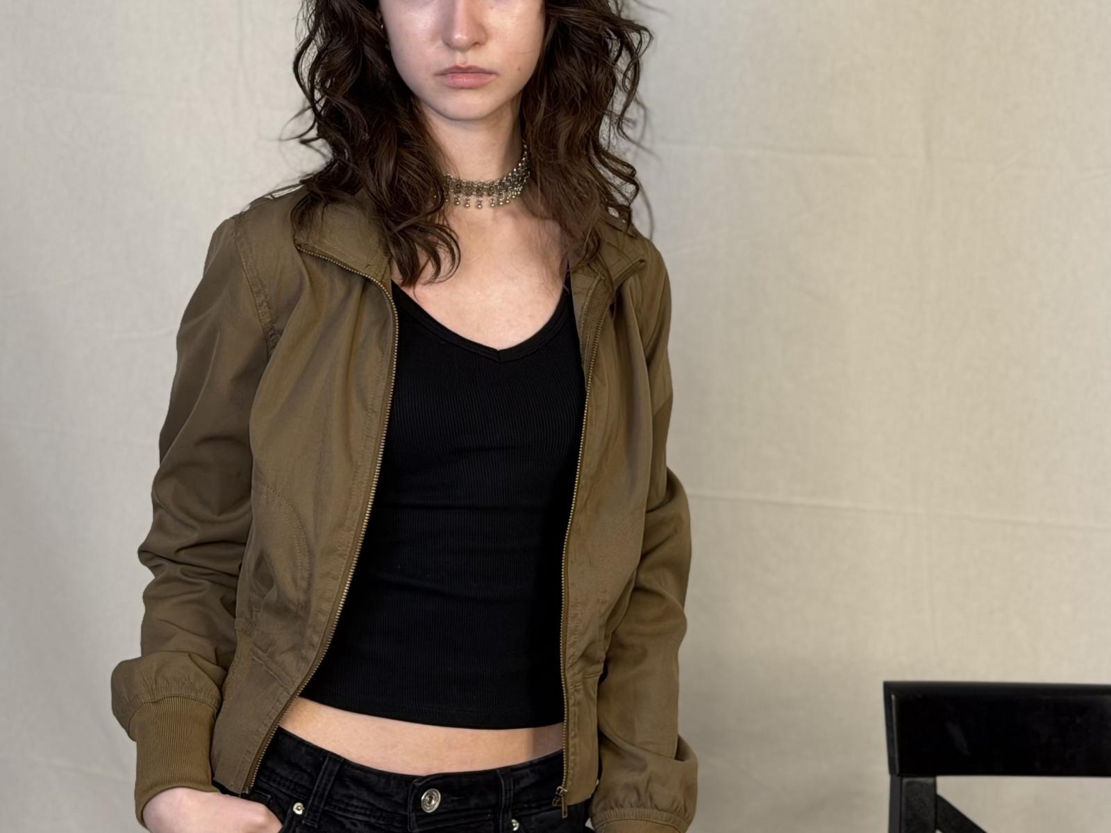 Jaclyn Palczewski, com cabelo castanho ondulado, vestindo uma jaqueta verde-oliva sobre uma cropped preta, mostrando suas nádegas planas em jeans escuros, e um colar choker..