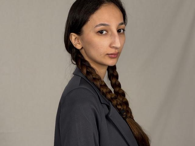 EarleenDalbeck has long brown braids, a serious face, and is wearing a dark gray shirt. She's looking over her shoulder with her brown eyes.