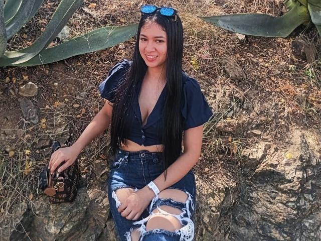 AnastasiaRouge with long black hair, blue eyes, small tits, wearing a navy crop top, ripped jeans, sunglasses on head, holding a black bag, sitting on rocks, smiling.