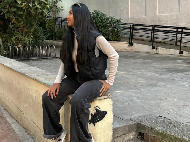 AnaRome sitting on a concrete ledge, wearing a black vest, beige long-sleeve shirt, blue jeans, and white sneakers with black straps, looking to the side.