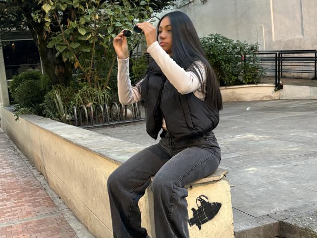 AnaRome with long black hair, wearing a black vest over a white long-sleeve shirt, and dark jeans, sits on a concrete block, holding a phone to her ear.