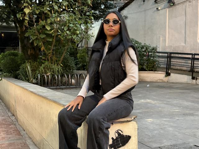 AnaRome, dark-haired woman, wearing sunglasses, black vest, beige long-sleeve shirt, gray pants, sitting on concrete wall, black shoes on ground, green plants, white wall in background.