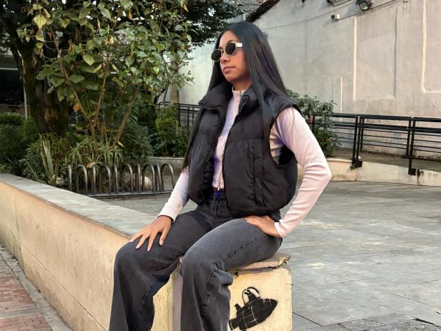 AnaRome, with long black hair and sunglasses, sits on a concrete block, wearing a black vest, white shirt, and flared jeans, hands on her knees, black bag on the ground.