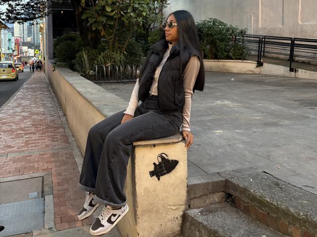 AnaRome sitting on a concrete wall, wearing a black vest, beige shirt, dark jeans, and white sneakers with black "C" logos. Black handbag on wall, sunglasses on face.