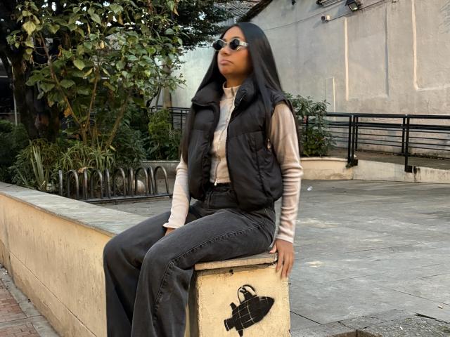 AnaRome with dark skin, long black hair, wearing sunglasses, a black vest, beige shirt, and black jeans, sitting on a concrete block, hand on knee, casual urban look.