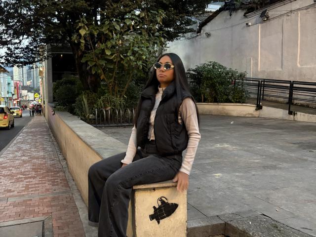 AnaRome with black hair, wearing sunglasses, a black vest, beige long-sleeve shirt, and black pants, sitting on a concrete block, holding a black purse, street background.