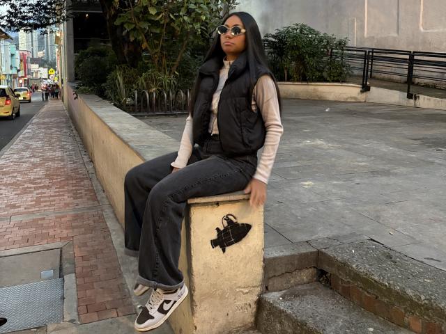 AnaRome with black hood, glasses, beige shirt, black vest, and black jeans, sitting on concrete ledge, white and black sneakers, black handbag on ledge, urban street background.
