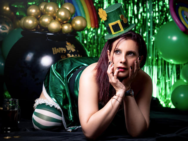 AdaMclain con un costume irlandese verde, calze a righe alte fino alla coscia, un cappello a cilindro verde e un trifoglio, su un tavolo nero, con le mani sul viso, che guarda verso l'alto, con palloncini verdi e dorati sullo sfondo..