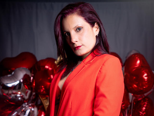 AdaMclain mit roten Haaren, die einen roten Blazer trägt und mit einem leichten Lächeln in die Kamera schaut, umgeben von glänzenden, herzförmigen Luftballons in Rot und Silber. Ihre Brüste sind teilweise sichtbar..