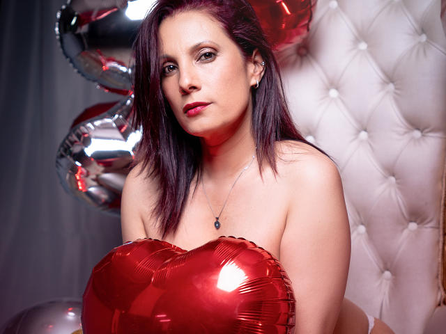 AdaMclain, com seios vermelhos e brilhantes em formato de coração, cabelo roxo escuro, batom vermelho e um colar, segurando balões, em frente a uma parede branca com textura..