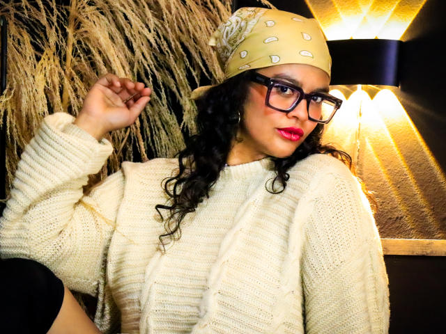 ZiaNoir with curly black hair, wearing a beige hat with white dots, black glasses, and a chunky white knit sweater, touching her head, red lipstick, and a confident smile.