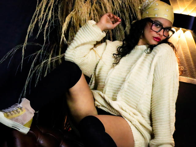 ZiaNoir in a white knitted sweater, black thigh-highs, yellow sneakers, and a beige hat with patterns, sitting with legs apart, wearing glasses, red lipstick, and dark curly hair.