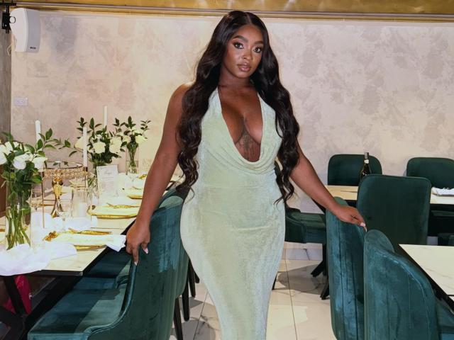 Nicoleth69 with dark skin, long wavy black hair, green halter dress with deep cleavage, showing her big boobs and curvy ass, standing in a restaurant with green chairs.