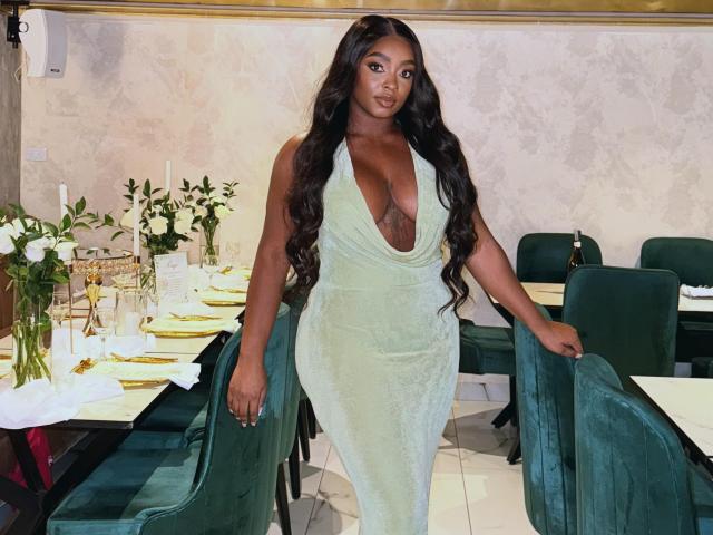 Nicoleth69, avec une peau foncée, de longs cheveux noirs ondulés, portant une robe en velours vert avec un décolleté plongeant, montrant ses grandes seins, se tient dans un restaurant, tenant une chaise verte..
