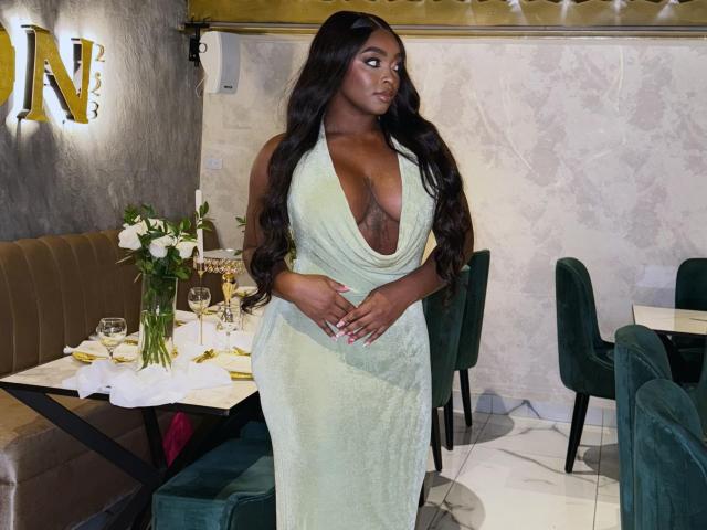 Nicoleth69, avec ses cheveux brun foncé, porte une robe vert foncé et profonde qui dévoile sa poitrine volumineuse et brillante, et se tient dans un restaurant élégant avec des chaises vertes et des fleurs blanches..
